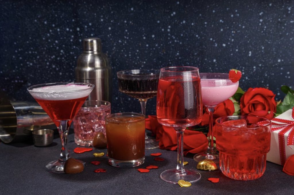 A series of red and pink cocktails with flowers and chocolates setting the scene around them. The background is blue-black with specks of white.