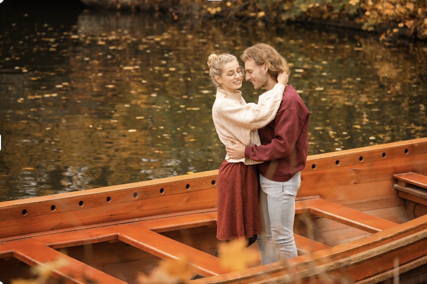 A happy couple holding each other while standing in a boat in the midst of autumn.