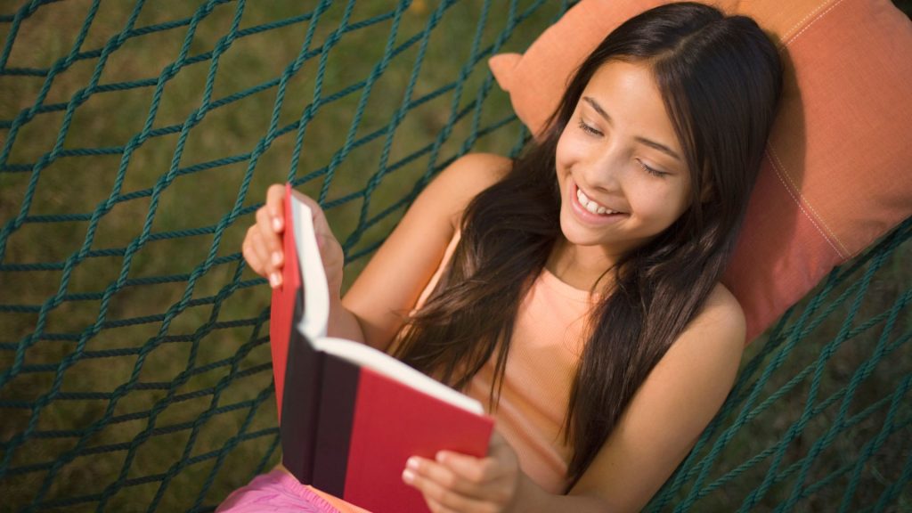 A young girl with light brown skin and long, dark brown hair lies in a hammock and happily reads a book.