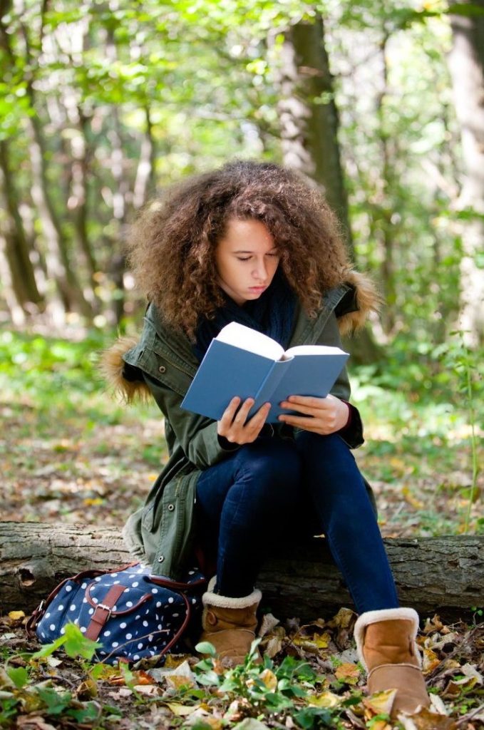 A girl with light brown skin and curly light brown hair sits on a log in the woods and reads.