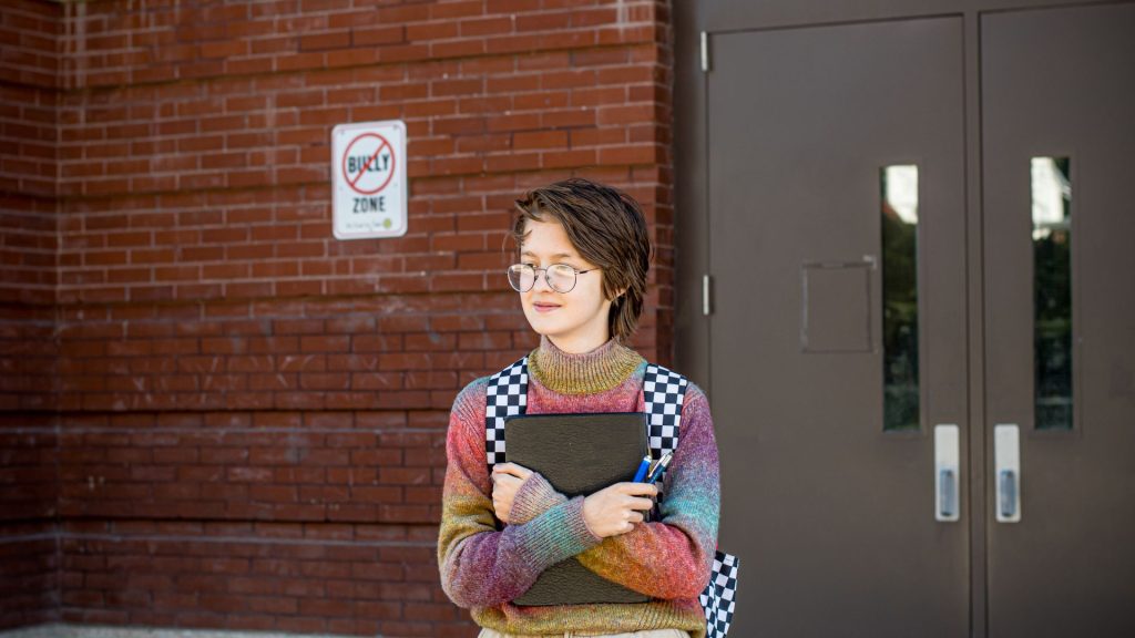 A white youth with short brown hair, glasses, and a colorful sweater stands outside a school. They are looking away and are holding a book against their chest.