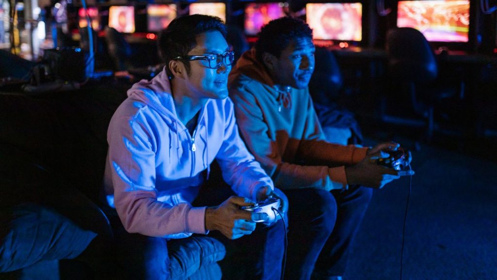 Two men sit in a dark room full of lit computers in the background. They face a screen off camera that projects blue light on their faces. Both men hold video game controllers.