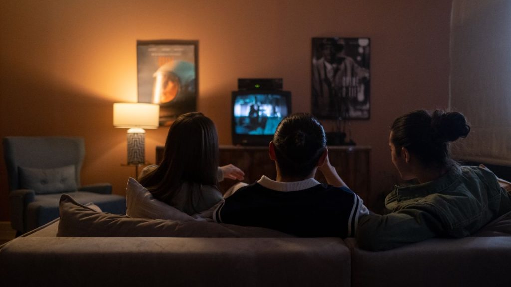 Three friends sit on a couch in a dimly lit room facing a small square TV. Two posters are on the wall and a lamp is lit in the left corner beside a gray armchair.
