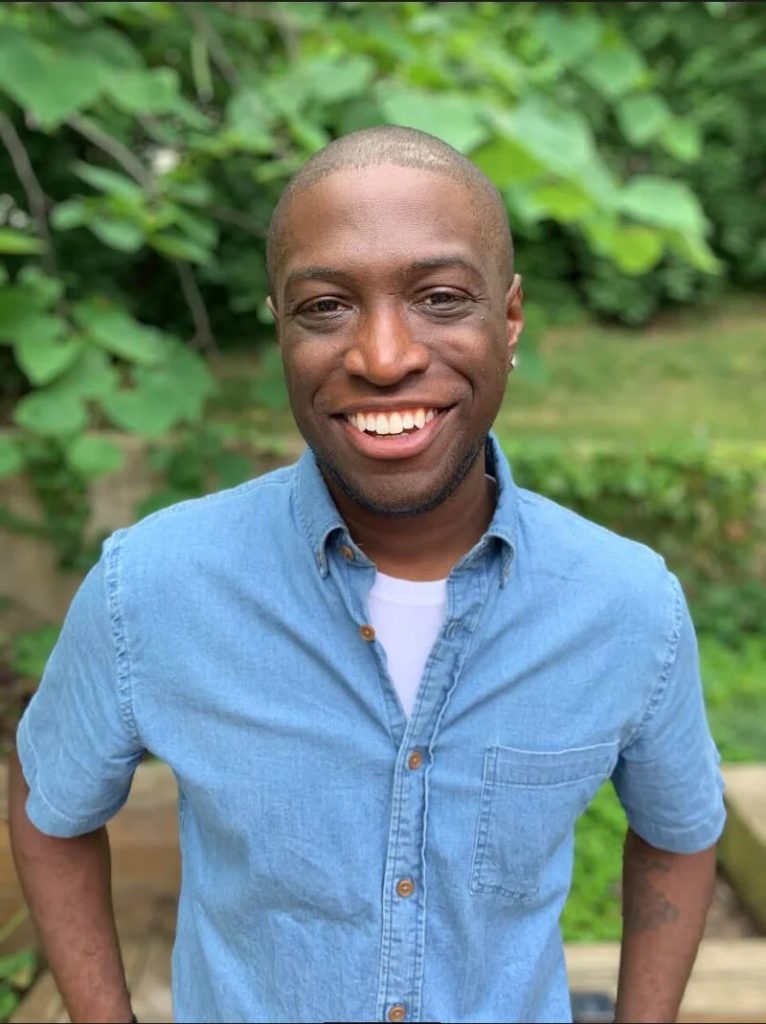 Vladimir Sainte wears a blue button down shirt and smiles in front of green foliage.