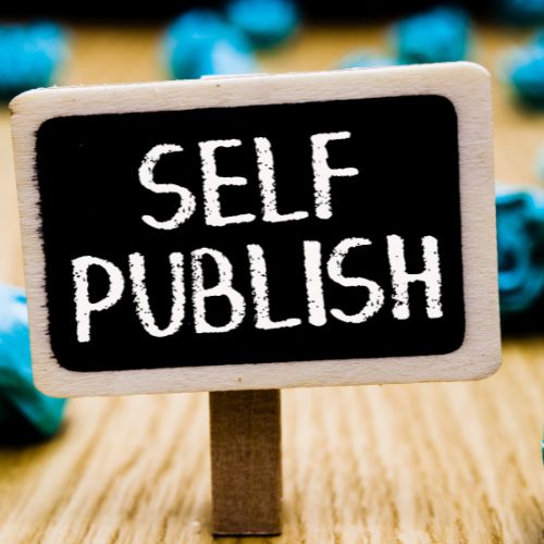 self publish written on a mini rectangle popsicle stick sitting on a piece of wood with blue-shaped balls scattered in the background