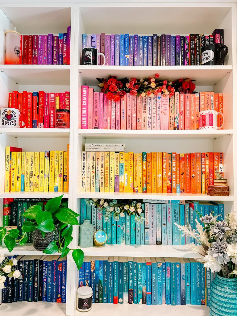 white bookshelves with rainbow color coded books. Coffee mugs, flowers, and other small decor pieces scattered throughout