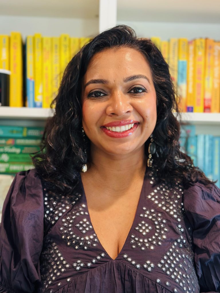 portrait of bookstagrammer Sarah smiling with a white beaded purple top against a bookshelf of yellow, green, and blue books