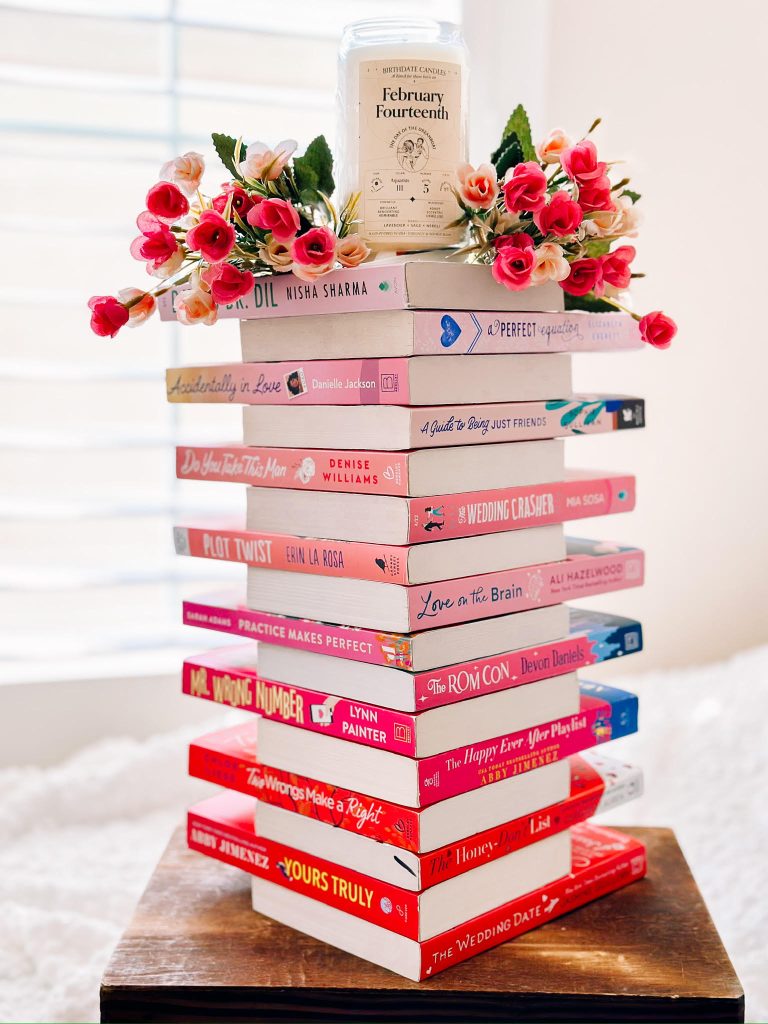 pink and red themed books stacked in a tower with a candle and flowers on top. set on wood stand on top of white bed sheets and a tan wall