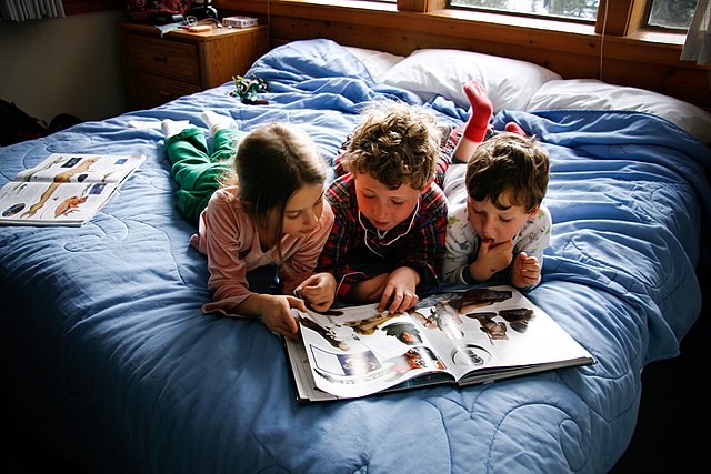 A picture with three young children laying on a bed with a book in front of them.