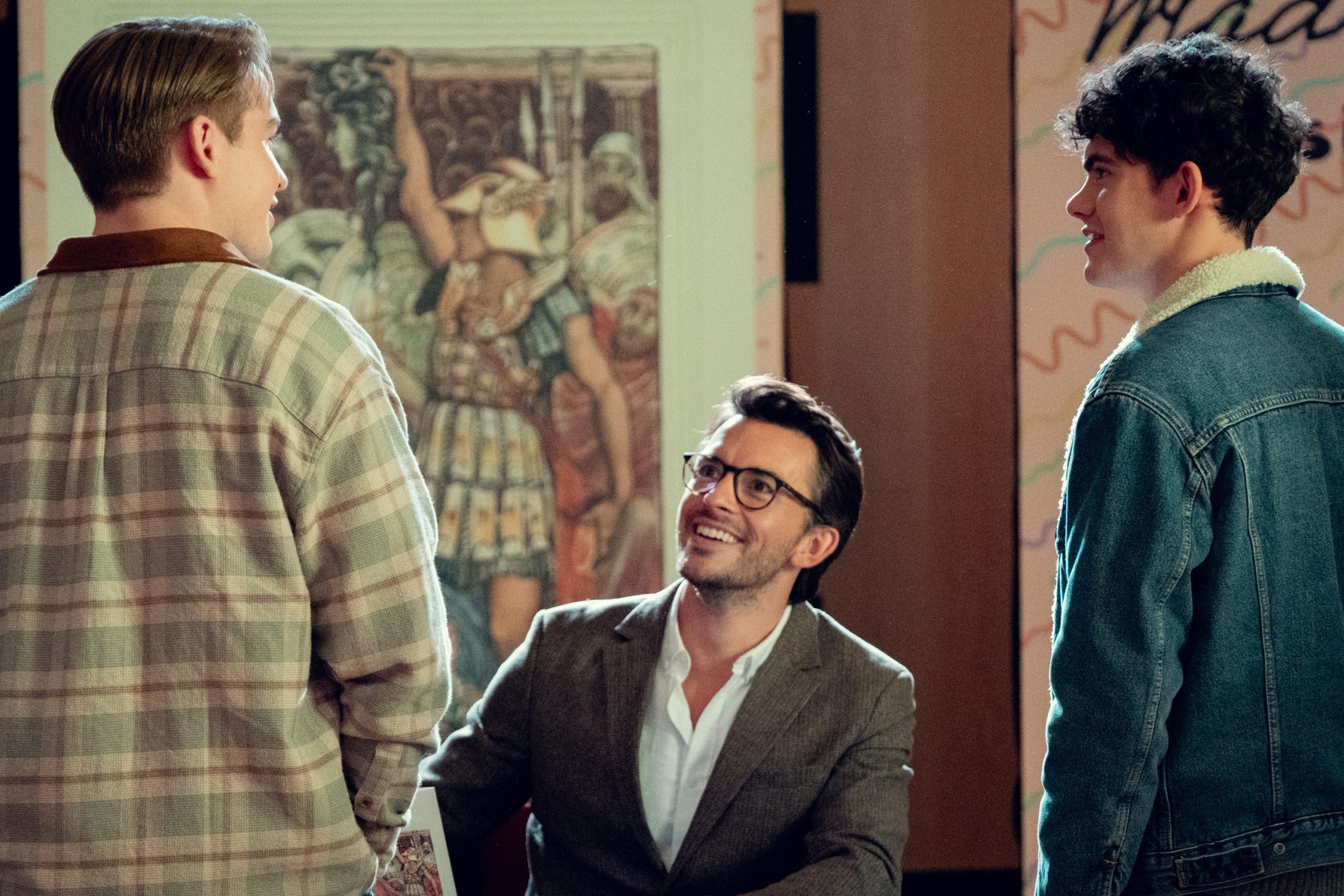 A picture of Jonathan Bailey in the Netflix show, Heartstopper. He's wearing glasses and a suit, smiling and looking at Nick, the main character of Heartstopper. Standing to the right is Charlie, the other main character of the show.