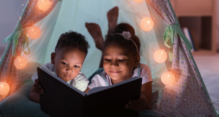 Two little kids, a boy and a girl, laying down reading in a tent adorned with lights, with a book between the two of them