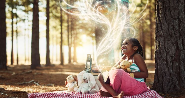 A little girl sitting on a picnic blanket in a forest looking at white, magical wisps emerging from her book and holding a teddy bear