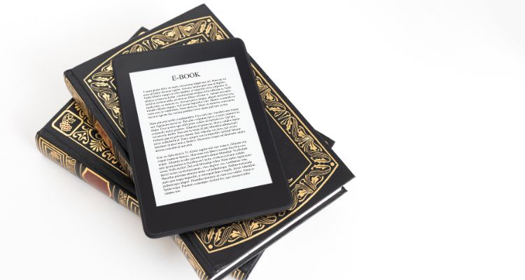 A stack of two books rests against a white background. On top of the books is a black e-reader. The e-reader shows some texts with the title "E-Book" on top.
