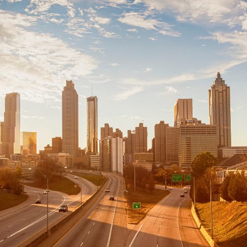A view of the Atlanta skyline during sunset. Three lines of traffic lead in and out of the skyscrapers with a few cars on the road.