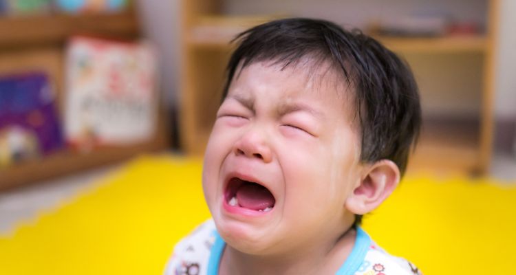 A small child is crying with his mouth open and eyes closed. In the background is a yellow floor cover and some shelves.