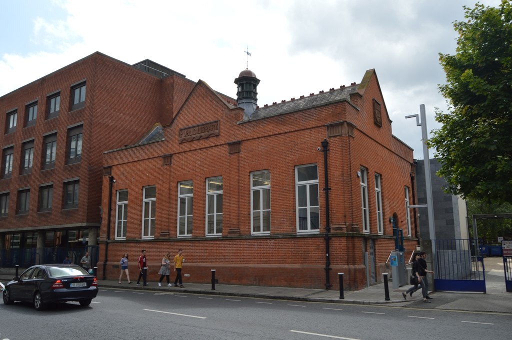 A historic orange brick building. There is a peaked roof with a small central turret and weathervane.