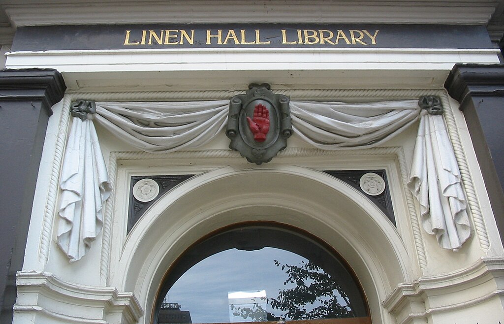 A building's entrance. Above the curved archway is white stone carved to look like curtains. In the center is an olive green crest with a red hand. Linen Hall Library is in gold lettering.