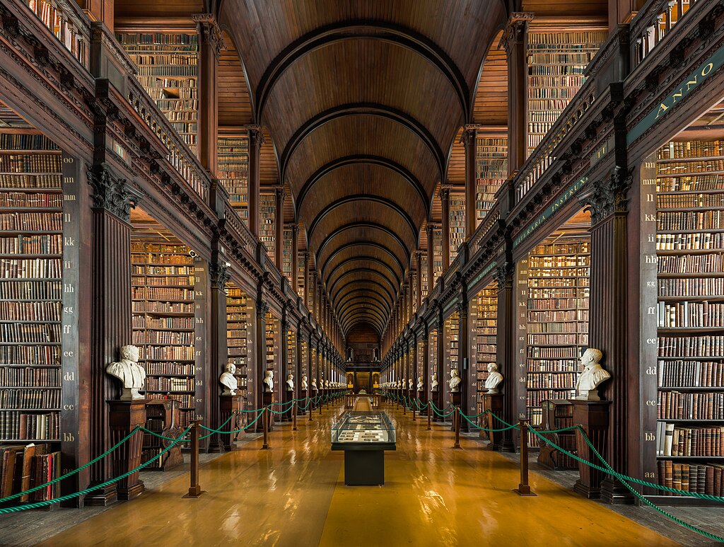 A long hallway with a curved wooden ceiling. Tall wooden bookshelves rise to a second story balcony. At the end of each aisle is a white bust. Green rope partitions off the shelves. Display cases are in the center of the hallway.