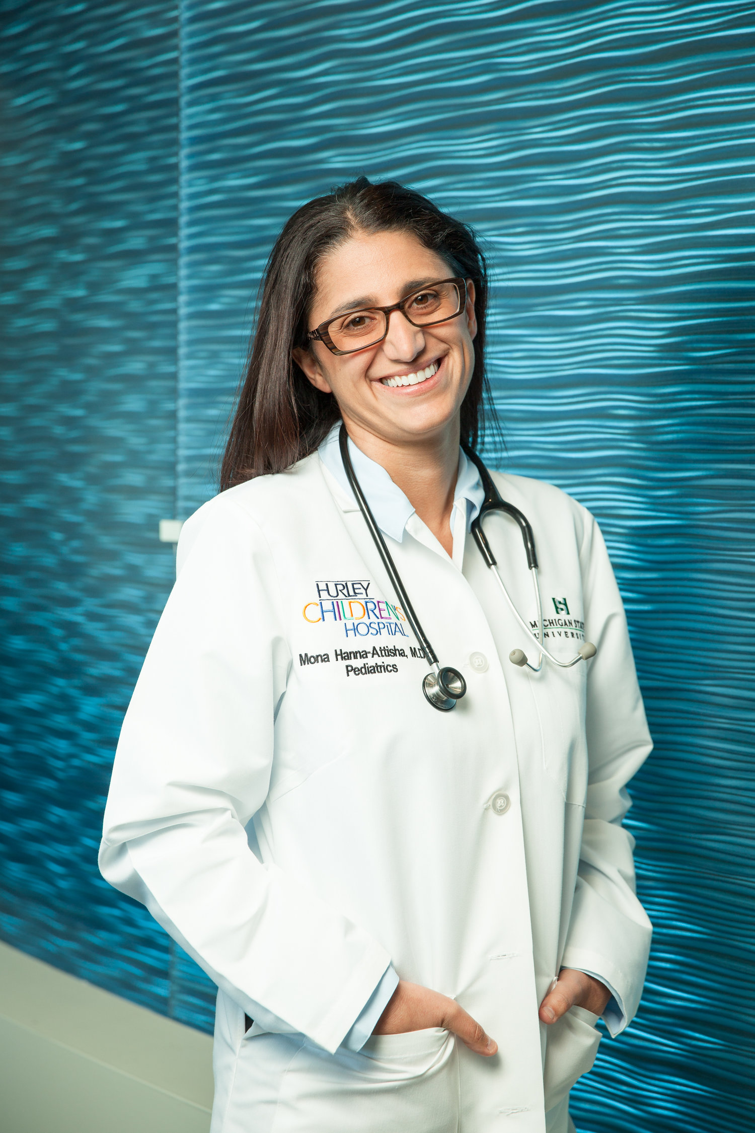 Author Mona Hanna-Attisha smiles wearing a lab coat and a stethoscope against a bright, blue-green background.