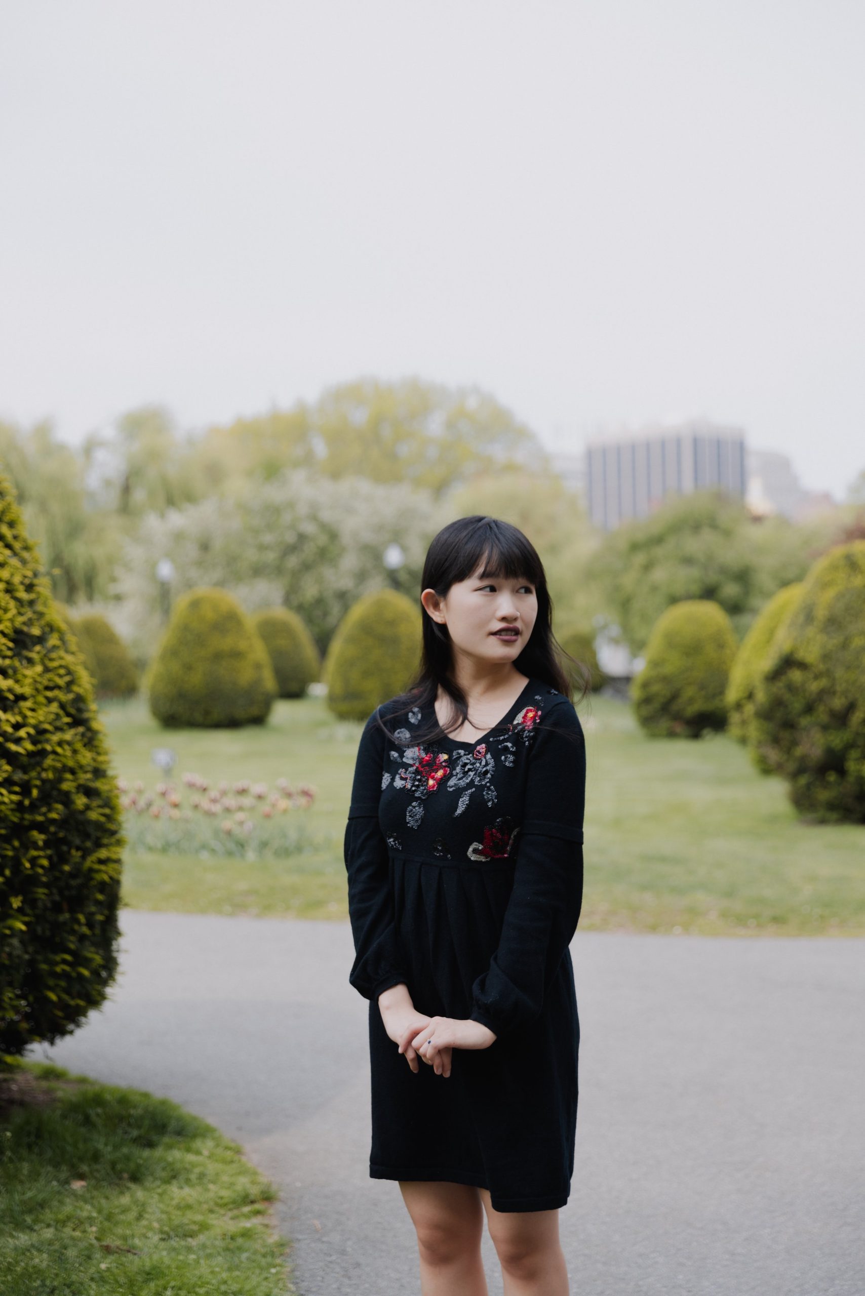 Author R.K. stands in a garden, wears a black dress with floral embellishments, and looks toward the left.