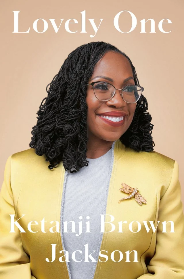 Ketanji Brown Jackson in a yellow blazer with a dragonfly broach and a beige background.