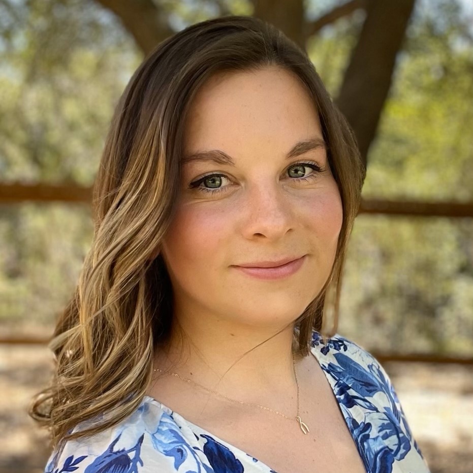 A smiling light-skinned woman with long, straight brown hair with blonde highlights, wearing a blue and white floral blouse and standing outside.