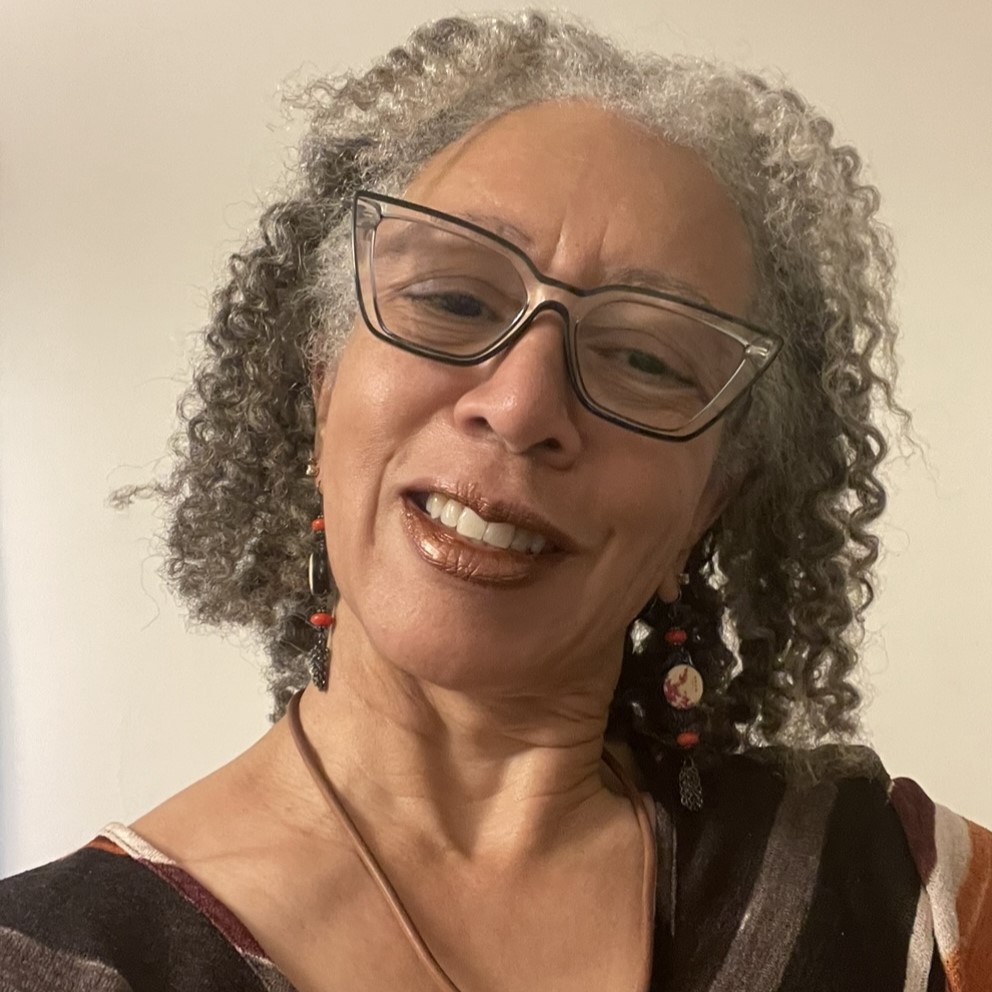 A smiling brown-skinned woman with short, curly gray hair and wearing glasses standing indoors.