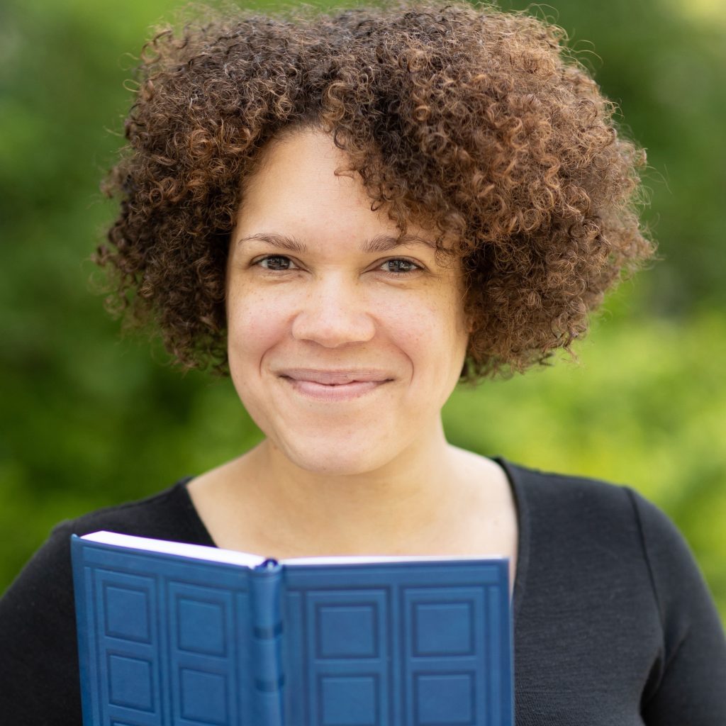 A smiling brown-skinned woman with short curly brain hair stands in a green outdoor area holding a blue book open.