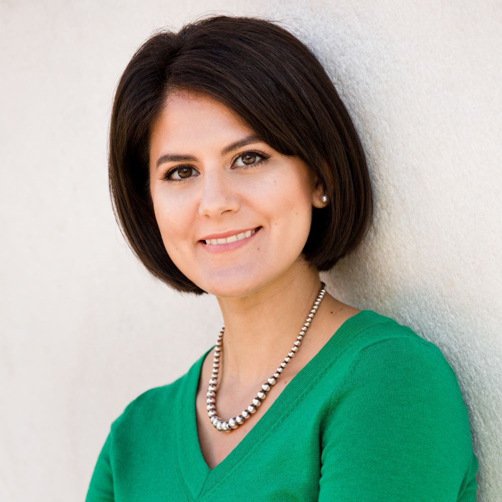 Emma Otheguy, a smiling light-skinned woman with short, straight dark hair wearing a silver beaded necklace and green sweater leans against a white wall.