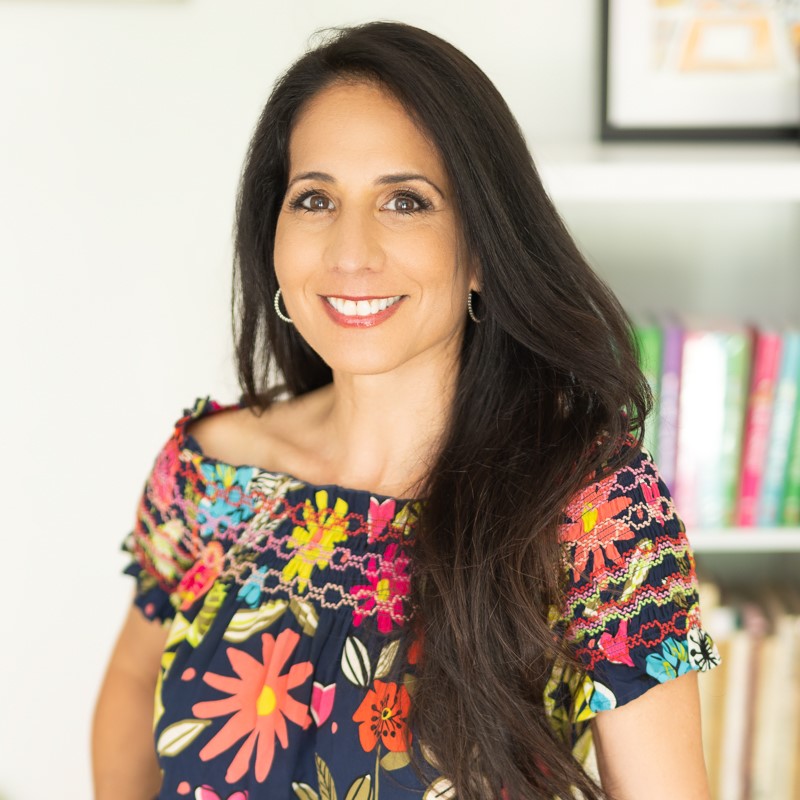 A smiling tan-skinned woman with long, straight dark hair wearing a floral blouse in front of a bookshelf with colorful books.