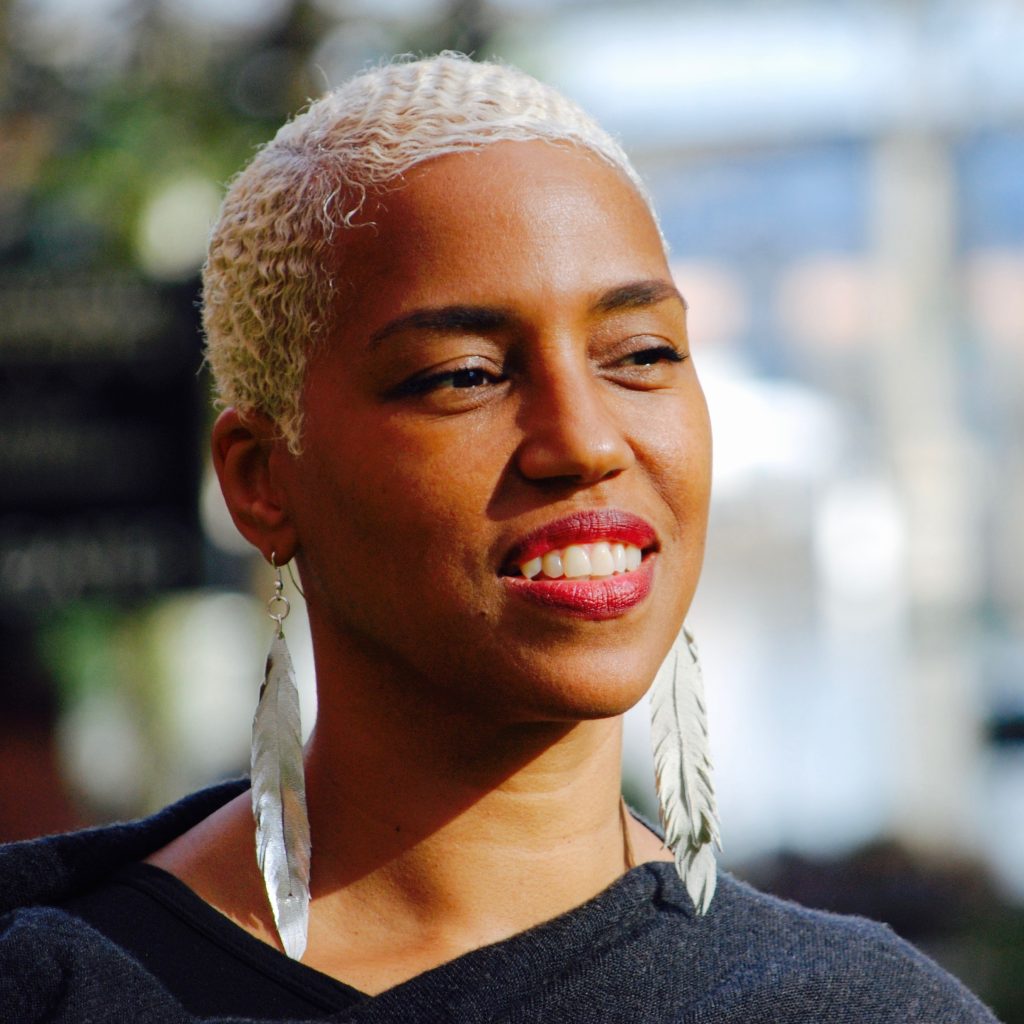 A smiling brown-skinned woman with short blonde hair and long feathered earrings stands outside.