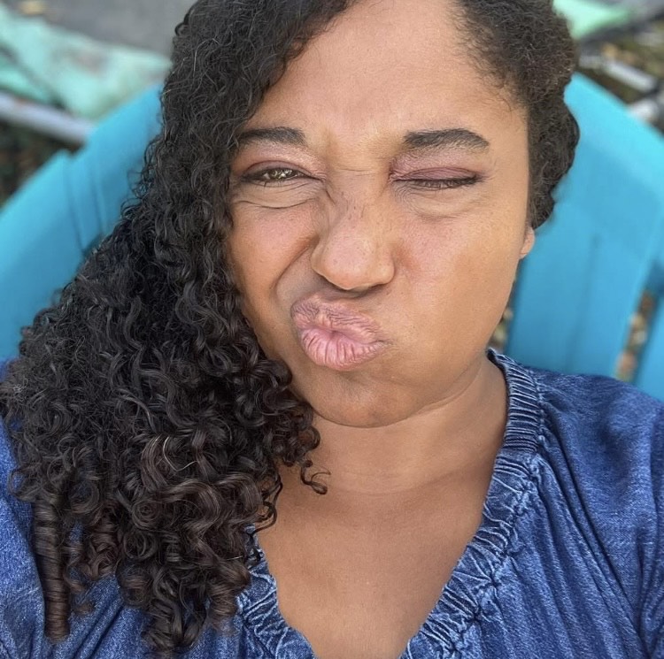 A brown-skinned woman with long, dark curly hair winks and makes a kissy face while sitting on a blue chair.
