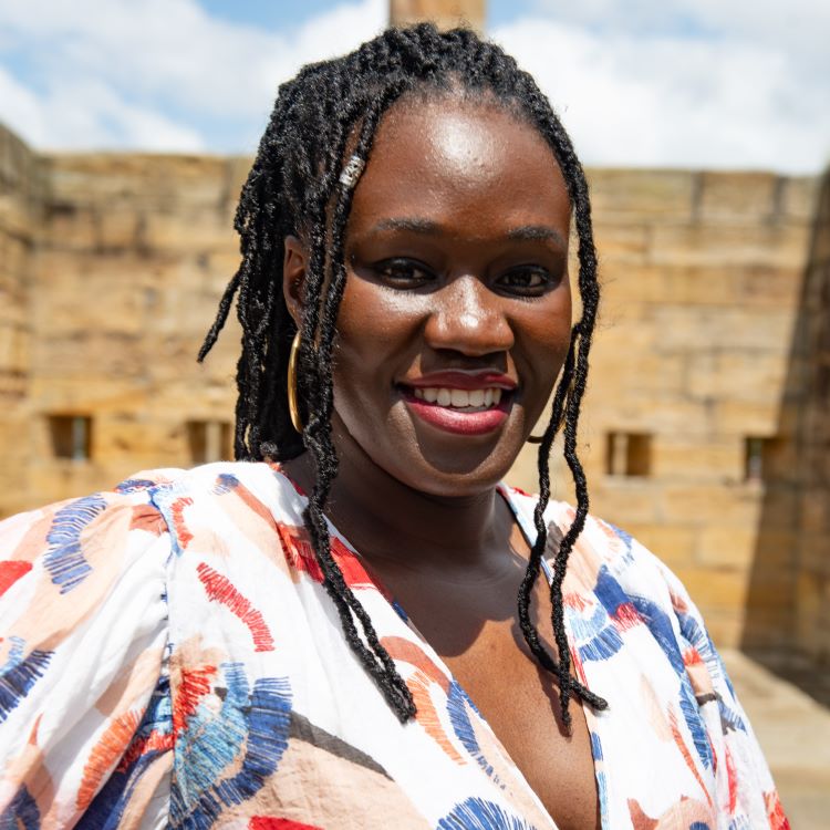 A smiling brown-skinned woman with long hair in minilocs wearing a patterned blouse and standing outdoors.