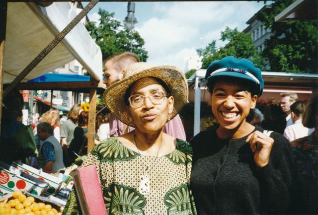 Audre Lorde dressed casually in green and tan and wearing a hat as she poses with a friend.