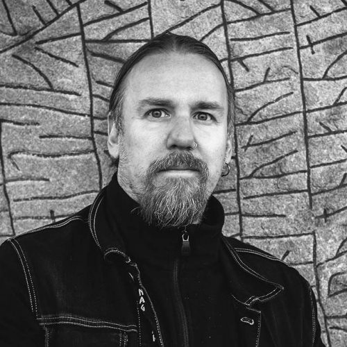 Portrait of Tommy Kuusela. He is wearing a black jacket and standing against a rune stone.