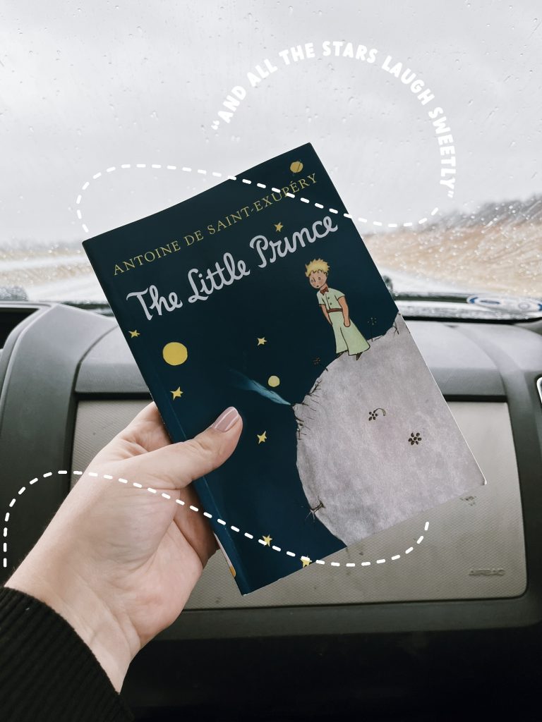 Kenzie G's arm holding up a copy of The Little Prince by Antoine de Saint-Exupery in a car. A white dashed trail swirls around her hand, leading to the words "And all the stars laugh sweetly" written in white all-caps.