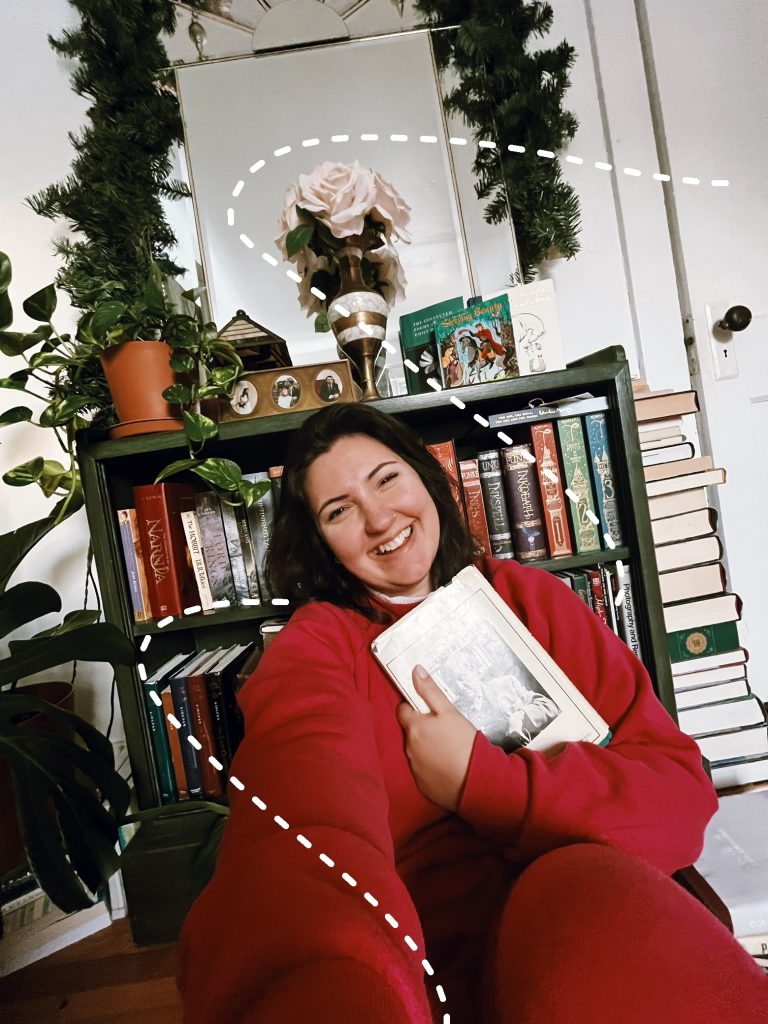 Kenzie G seated in front of a bookshelf, holding up a book and smiling at the viewer. A dashed white trail swirls behind her.