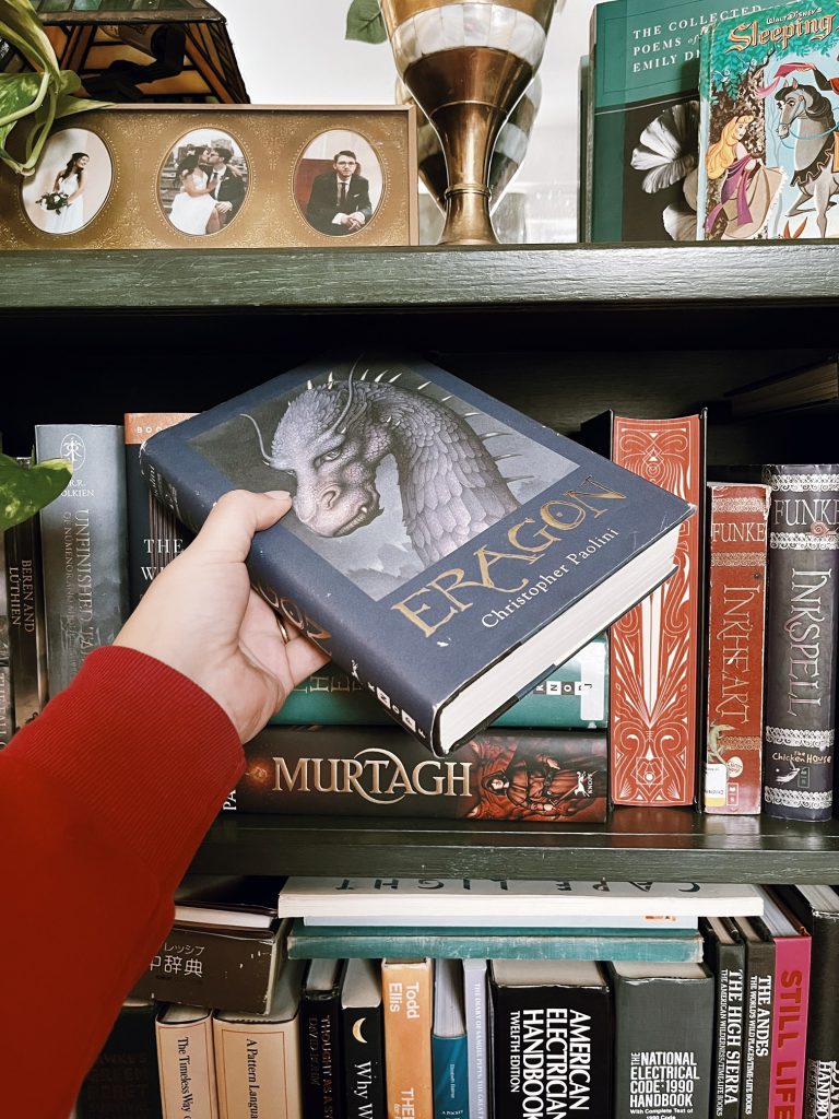 Kenzie G's arm holding up a copy of Eragon by Christopher Paolini in front of a bookshelf.