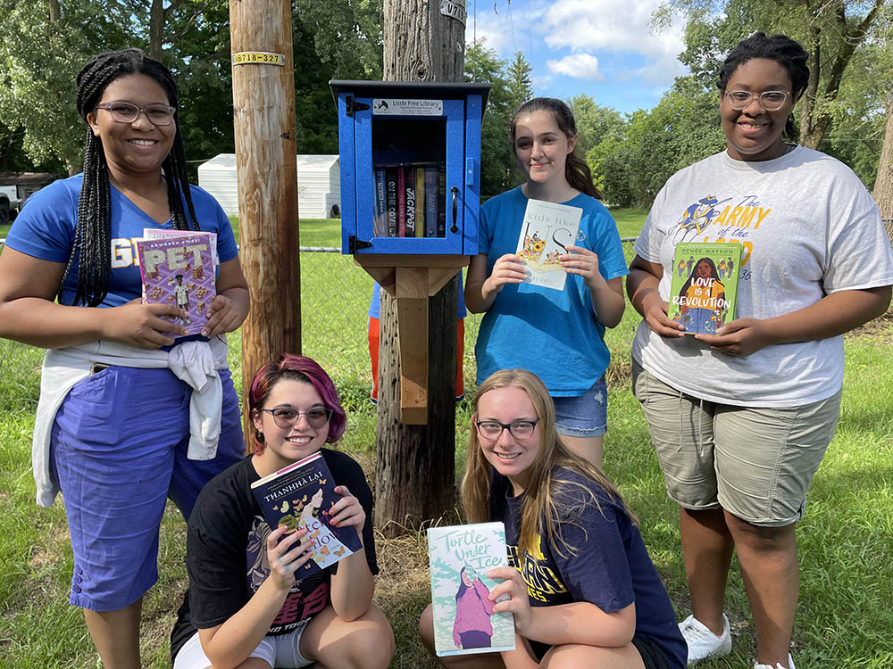 A dark blue little library is attached to a telephone pole. Five teens pose around it with books in their hands. Grass and trees are in the background.