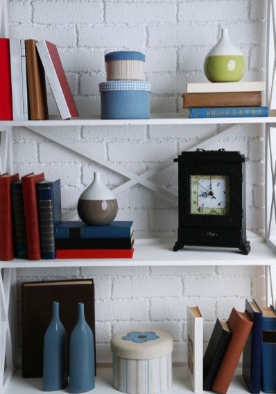 A small bookcase with small stacks of books, little stacked containers, a clock, two bottles, and two small containers.