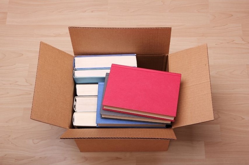 Eight books stacked in a cardboard box sitting on a light wooden floor.