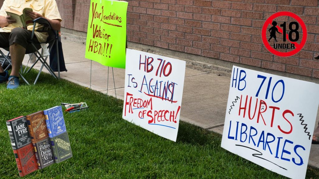 Protest signs fighting back against the HB 710 ban stand in a row. The advisory symbol of under 18s banned is pictured in the top right. A stack of classic literature rests in the lower left side of the image.