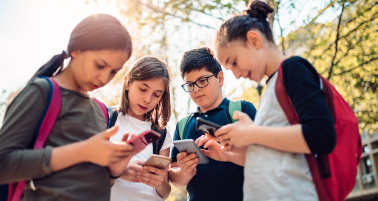 A group of children wearing backpacks and standing in a semicircle stare down at smartphones.