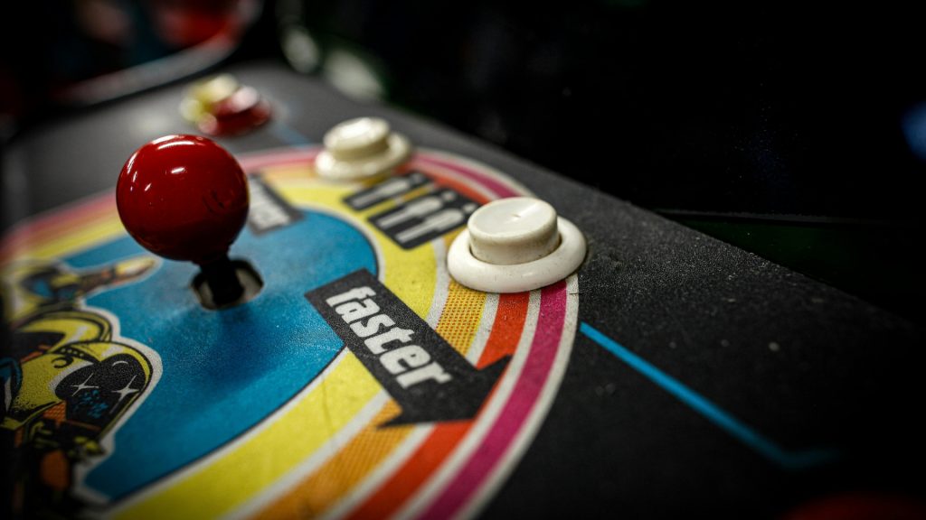 A close-up of a retro arcade game. A black background with white, yellow, and red buttons and a red joystick. A multi-colored circle design in the center.