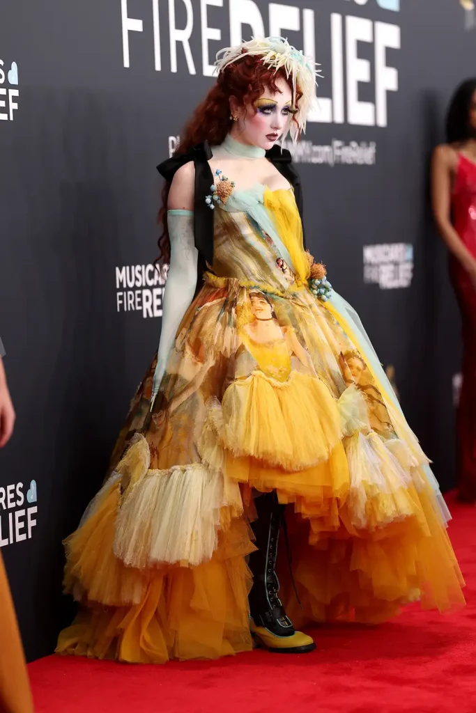 Chappell Roan wears a striking yellow and multicolored gown featuring artistic prints, layered ruffles, and floral embellishments on the red carpet. The look is completed with a feathered headpiece, long gloves, and bold black-and-yellow boots.