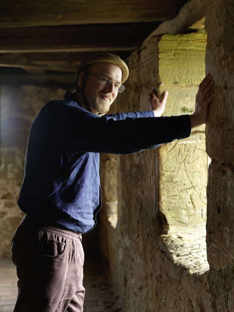 A photo of author Robert Thier in a building that looks similar to a bunker. Wearing a long-sleeve blue button up and brown pants wearing glasses.