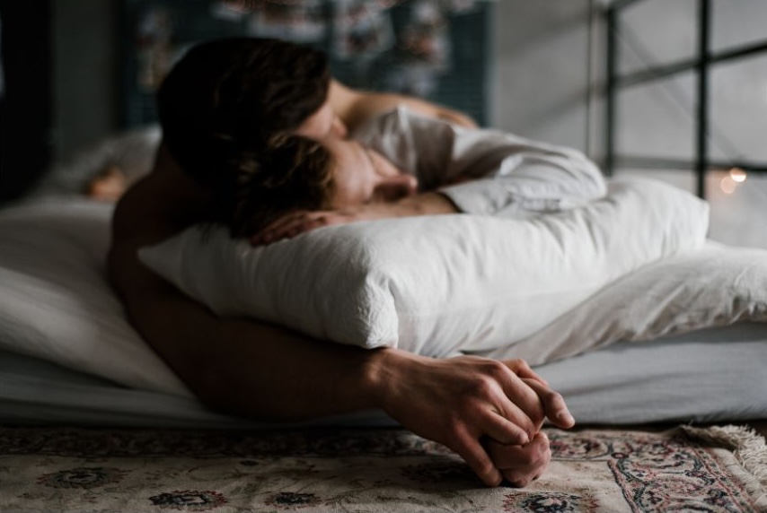 A photo of a couple cuddling and holding hands in bed.