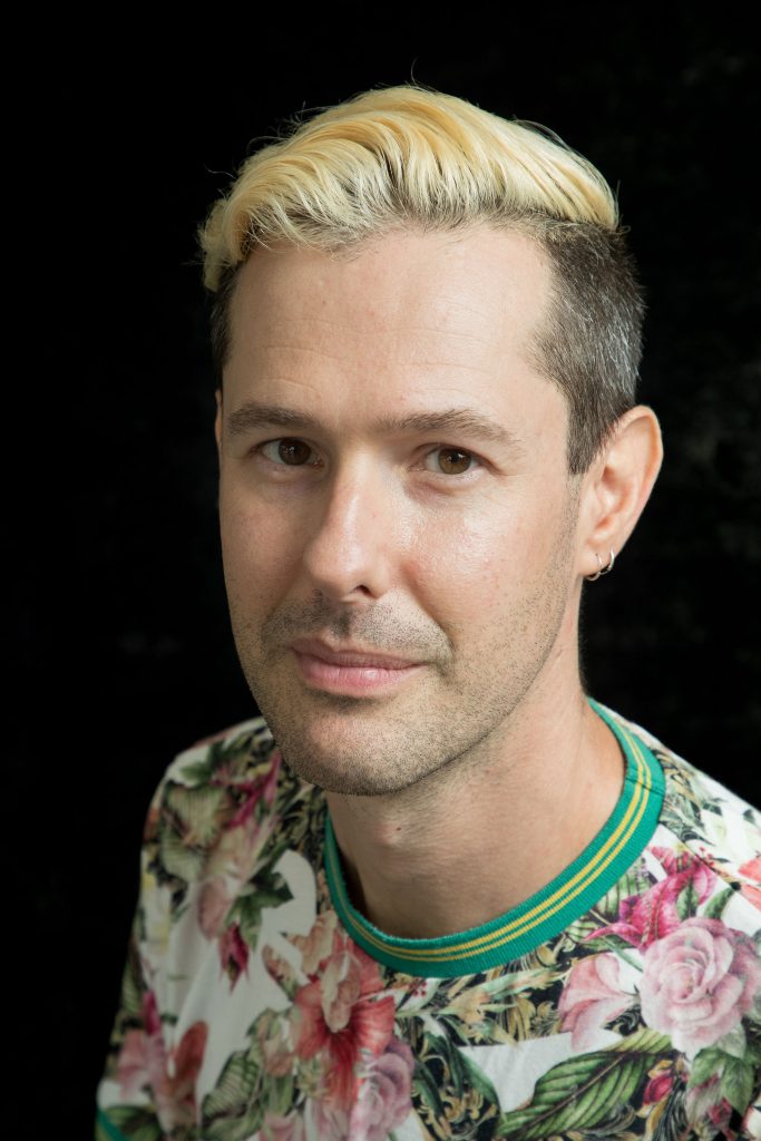 A photo of author Liam Livings smiling with bleached short hair, earrings, and wearing a floral t-shirt.