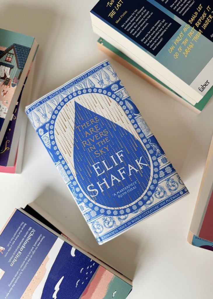 Image of a few books laid out. Four books are half in frame, with the center of the image being There Are Rivers In The Sky by Elif Shafak. It is a blue book with a teardrop shape in the middle.
