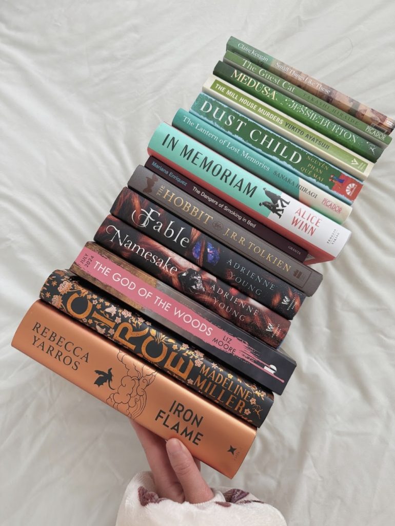 A stack of books laid spine facing upwards on a sheet. The books get smaller the higher on the stack they get. The focus of the image is on the first two books: Circe by Madeline Miller and Iron Flame by Rebecca Tarros.